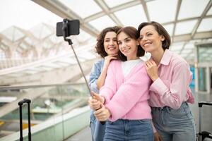 Three friends pose for a selfie with joyful expressions while standing in a bright airport terminal, surrounded by contemporary architecture and luggage. photo