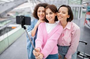 Three young women smile and pose for a selfie using a selfie stick in a bright, modern building. They are enjoying their time together and capturing the moment. photo