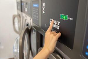A person selects the wash cycle on a self-service laundry machine in a brightly lit laundromat. The machine displays settings and remaining time for the laundry process. photo