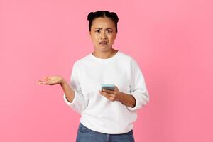 Asian woman with a puzzled expression holds her smartphone and gestures with her hand. She appears to be questioning something as she stands in front of a vibrant pink background. photo
