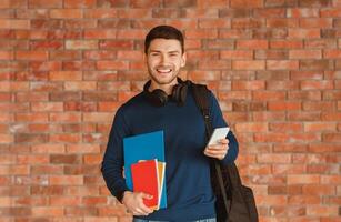 Modern Student. Happy Guy With Backpack Holding Smartphone And Notebooks Smiling To Camera Standing Over Brick Wall Background Indoor photo