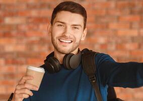 Positive Guy Making Selfie Smiling To Camera And Holding Paper Coffee Cup Standing Over Brick Wall Indoor photo