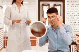 Handsome young man choosing new eyewear at optical shop with the help of lady specialist, looking in mirror photo
