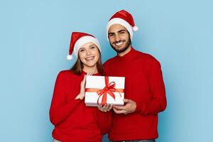 A cheerful couple in matching red sweaters and Santa hats stands against a light blue background, smiling and presenting a wrapped gift with a red ribbon, radiating holiday joy. photo