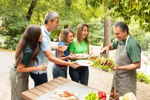 A group of five friends shares laughter and delicious food at a barbecue party in a vibrant garden. They eagerly await grilled meat while enjoying their meals and drinks. photo