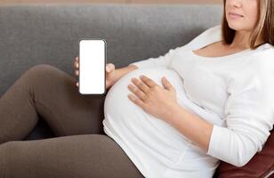 A pregnant woman sits comfortably on a couch, gently cradling her belly, holding smartphone with blank screen in her hand. The warm and inviting setting suggests a moment of peaceful anticipation. photo