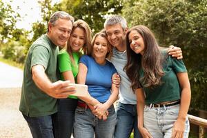A group of five friends stands close together outdoors, smiling and capturing a selfie on a phone. They are dressed casually, enjoying their time in the warm sunlight surrounded by greenery. photo