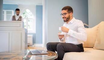 A man in a white shirt sips coffee and smiles while working on his laptop in a cozy lounge. A staff member can be seen in the background providing service, creating a relaxed atmosphere. photo