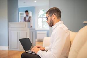 A man wearing glasses and a white shirt sits on a couch in a hotel lobby. He is working on a laptop. A person standing behind a desk in the background is out of focus. photo