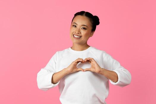 Asian woman is smiling while creating a heart shape with her hands in front of a bright pink backdrop. She showcases joy and affection in this vibrant setting. photo
