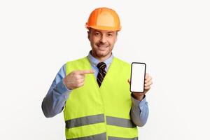 Construction Worker Showing Smartphone Blank Screen, Pointing Finger Recommending Building Services Standing In Studio On White Background. Mockup photo