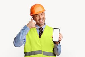 Builder Man Showing Phone Blank Screen Gesturing To Call Him Posing Over White Studio Background. Building Services Concept. Mockup photo
