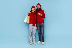 A happy couple poses together, showcasing colorful shopping bags and a gift card. They wear matching red sweaters and hats, smiling joyfully in a bright blue environment. photo