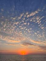 A stunning sunset over the ocean with a bright orange sun, casting light across the water and illuminating the scattered golden clouds in the expansive sky photo