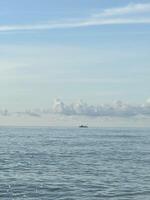 A small boat is seen far on the horizon, floating on a calm ocean under a bright and clear sky with a few scattered clouds. Peaceful maritime scene photo