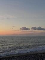 Soft waves gently break on a pebble beach as the sun sets, casting warm pastel colors across the sky and calm ocean. A tranquil and peaceful seaside scene photo