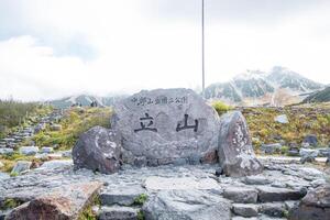 Murodo, Tateyama Kurobe Alpine Route, Japan, A stone monument with text Chubu Sangaku National Park Tateyama Murododaira 2450 in Japanese, a mountainous trail, surrounded by lush greenery photo