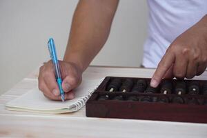 Close up man hand holds pen, write on paper and calculate on wooden abacus. Concept, ancient style calculation. Counting equipment. Calculator. Educational material. photo