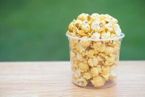Popcorn in glass bowl on wooden table against green background, made from seeds of maize or corn by baking or heating, mixed with salt, sugar and butter caramel. Concept, delicious snack. photo