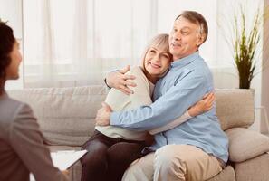 Psychotherapy. Happy senior couple sitting on couch and hugging, smiling female doctor listening, copyspace photo