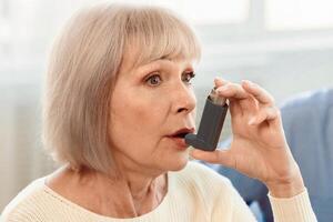 Senior Woman Using Asthma Inhaler For Allergies In Modern Clinic, Copy Space photo
