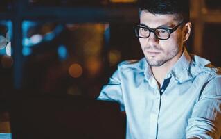 IT Specialist Guy At Laptop Working Coding Website Overnight, Wearing Eyeglasses Sitting At Workplace Indoor. Selective Focus photo
