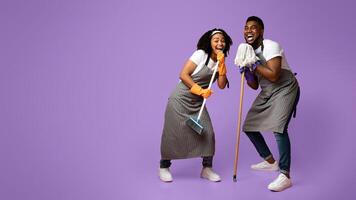 Domestic Band. Happy Black Couple Fooling Together While Cleaning Flat, Singing Into Mop And Broom, Standing On Pink Background In Studio, Free Space photo