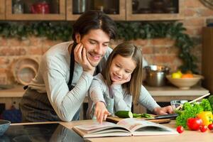 Caring young dad and little daughter searching recipe in culinary book, preparing healthy dinner together in kitchen, copy space photo