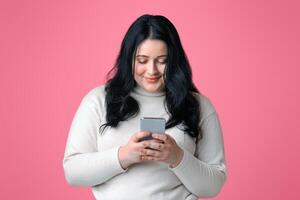 Smiling plump woman holding smartphone in hands, texting, reading message, or using new application, isolated on pink background with free space photo