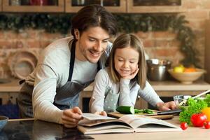 Smiling Little Girl And Her Dad Checking Ingredients In Coolinary Book While Preparing Healthy Lunch In Kitchen Together photo