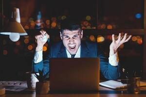 Angry Entrepreneur Screaming Working On Laptop And Crumpling Paper In Rage Sitting In Modern Office At Night. Business Failure Concept photo