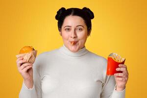 Not Counting Calories. Funny Fat Girl Eating Unhealthy Burger And French Fries Standing On Yellow Background In Studio, Empty Space photo