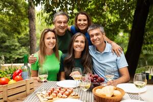 A group of five friends gathers around a table filled with wine and delicious food in a vibrant outdoor setting. Laughter and joy fill the air as they celebrate together. photo