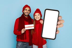 A cheerful couple dressed in red outfits holds a gift certificate while showcasing a blank smartphone against a light blue background. They exude joy and excitement in their cozy attire. photo