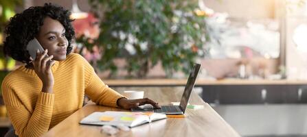 Young black businesswoman having conversation with clients on phone, working at cafe, using laptop, free space photo