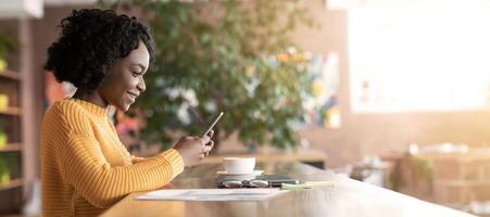 Pretty afro girl drinking coffee, working and using smartphone, cafe interior, copy space, side view photo