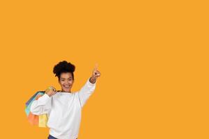 A joyful woman wearing a white sweatshirt holds multiple colorful shopping bags while pointing excitedly. The vibrant orange background adds to her cheerful expression as she shops. photo