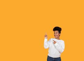 A young woman in a white sweatshirt joyfully points to the side, smiling brightly against an energetic orange backdrop, showcasing enthusiasm and positivity in her expression, copy space photo