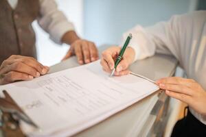 A person is signing a document with a green pen. The person's hand is holding the pen and their fingers are resting on the document. Another person is out of focus in the background. photo