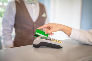 A customer is processing a payment at a retail counter, holding a green card near a card reader, while a staff member stands in a brown vest nearby, ready to assist. photo