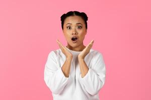 Asian young woman with two playful buns showcases a surprised expression while raising her hands in awe. The striking pink background enhances her joyful demeanor, capturing a moment of delight. photo