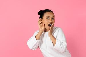 A young Asian woman with a joyful expression is shouting excitedly while standing in front of a pink backdrop. She has her hands raised near her mouth, conveying enthusiasm and joy. photo
