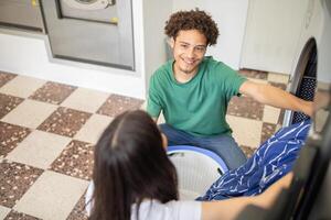 A young man smiles at a woman as they work together in a laundromat, loading clothes into a washing machine. The bright space features modern appliances and tiled flooring. photo
