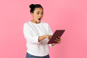 Asian woman with a shocked expression points at her tablet while wearing a white sweater, set against a vibrant pink backdrop. Her reaction suggests excitement or disbelief at what she sees. photo