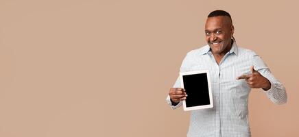 Smiling middle aged black man pointing finger at blank digital tablet screen, orange studio background, empty space photo