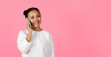 A cheerful Asian woman is engaged in a phone conversation, smiling brightly while wearing a light sweater. The vibrant pink backdrop enhances the joyful atmosphere of the moment, copy space photo