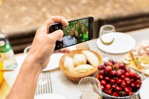 A hand holds a smartphone capturing a group selfie during a family gathering outdoors. Delicious food items like fruits and bread are displayed on the table, enhancing the cheerful atmosphere. photo