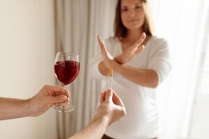 A woman with a serious expression is politely rejecting a glass of red wine offered to her. She holds a test stick in her other hand, indicating a significant decision. photo