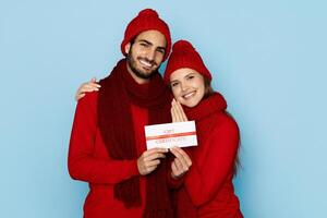 A joyful couple dressed in matching red winter attire holds a gift certificate, smiling warmly at each other against a light blue background, embodying the spirit of giving during the winter season. photo