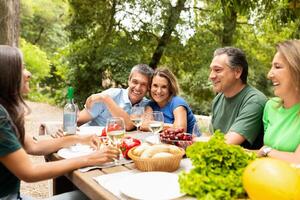 A group of five friends gather around a wooden table adorned with fresh food, laughing and sharing stories in a sunny park setting. Their joyful interaction highlights a vibrant summer day. photo